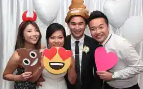 Wedding guests posing with fun emoji props including heart eyes, poop emoji hat, and heart sign in a photo booth
